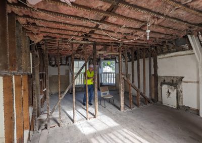 Image of someone in a hard hat from St IVes CLT in the Old Vicarage. The insulation and wires in the ceiling is exposed and the walls are not finished on the right side or middle partition of the room.