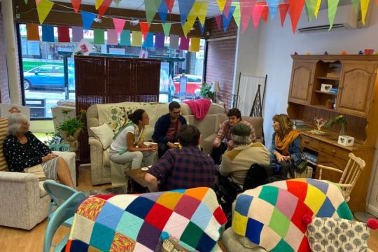 Image of people sitting in Hastings Commons CLT's common room. People are sat on sofas and armchairs. The room has bunting hanging from the ceiling and colourfully knittted blankets draped over the sofas. It looks cosy and lively