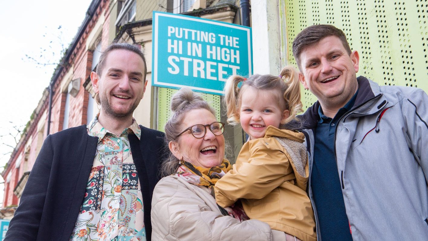 4 people stand in front of a row of terraced homes in front of a sign saying "Putting the Hi in highstreet". In the middle is a woman holding a toddler