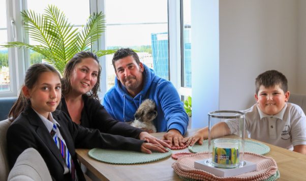 A family of four and their dog sitting in their new home around the table with their hands touching in the middle of the table