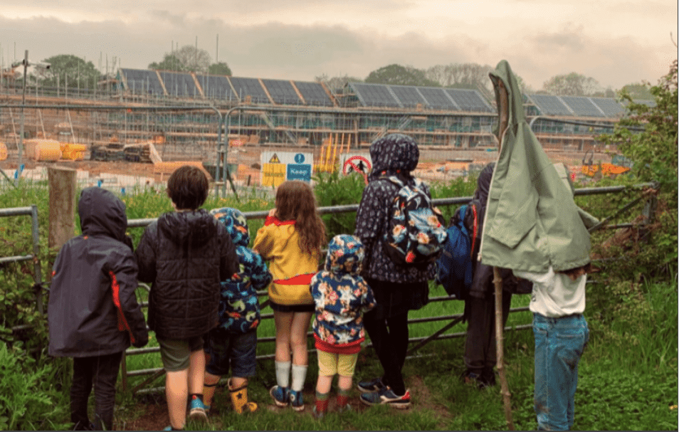 Young children in coats and wellies peeking over the fence onto the construction site for their new community led homes