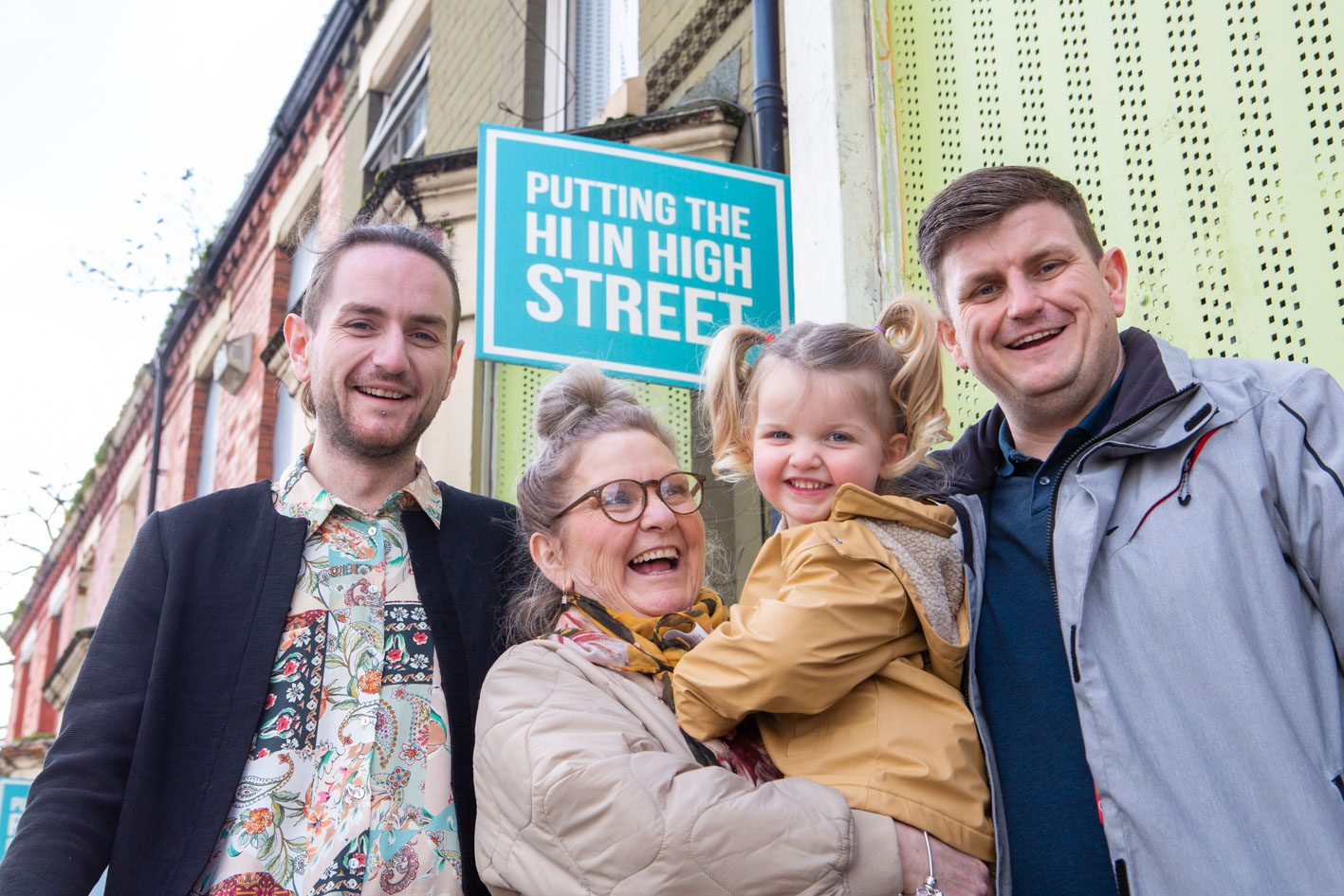 Home Baked CLT-1996 (2) Three adults and a toddler from Homebaked Community Land Trust, Anfield Liverpool stand in front of a sign saying "putting the high into highstreets". This is on a derelict terraced home that they are aiming to bring into their CLT's ownership